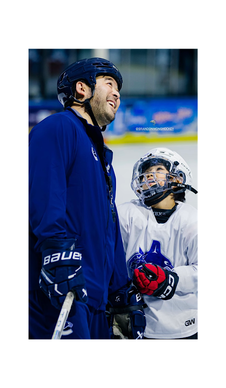 Two ice hockey players, one smiling and the other looking up, on the ice rink wearing helmets and gear.