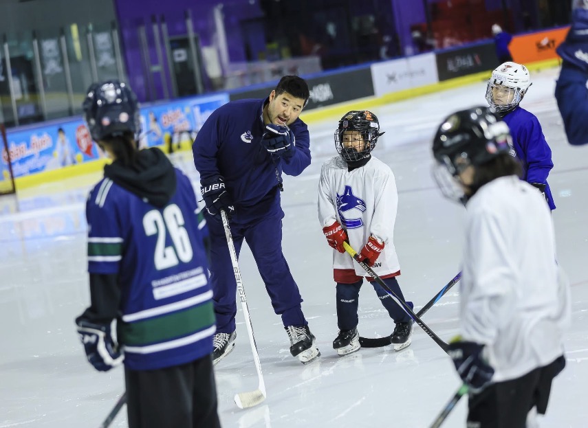 Hockey coach instructs young players on ice rink, children in hockey gear listen attentively.