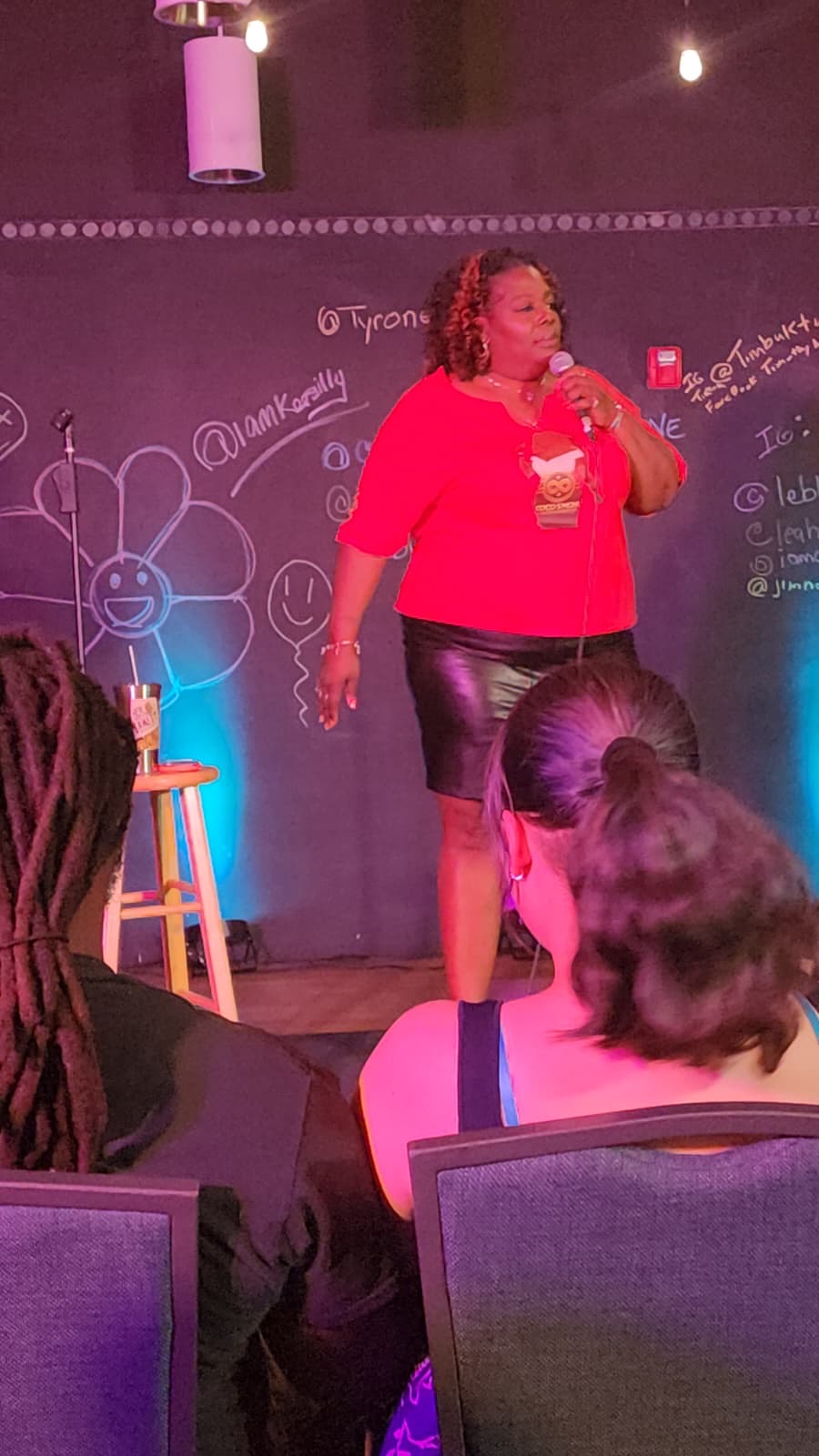 Woman standing on stage in front of a blackboard, speaking to an audience, with people seated in front.