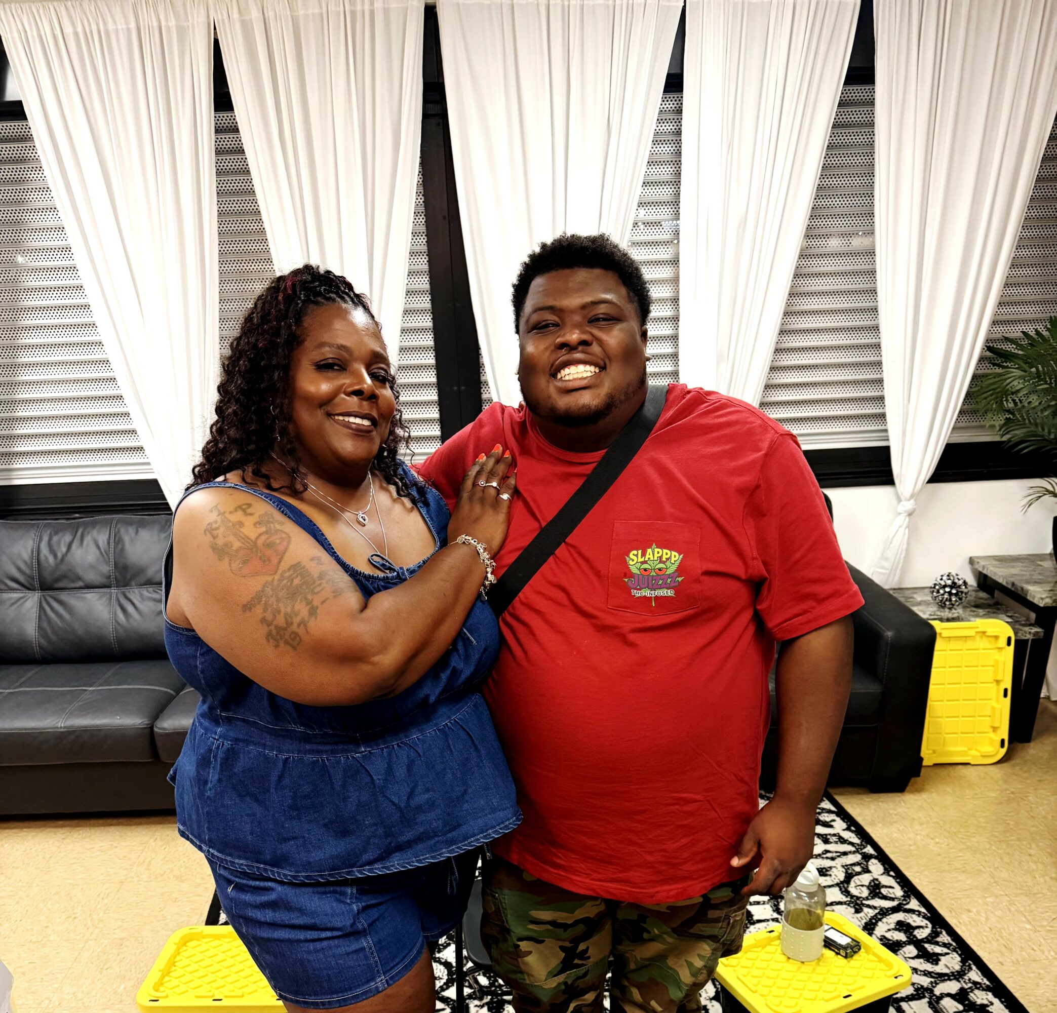 Two people smiling indoors, woman with curly hair and tattoos, man in red shirt, standing close together, curtains behind.
