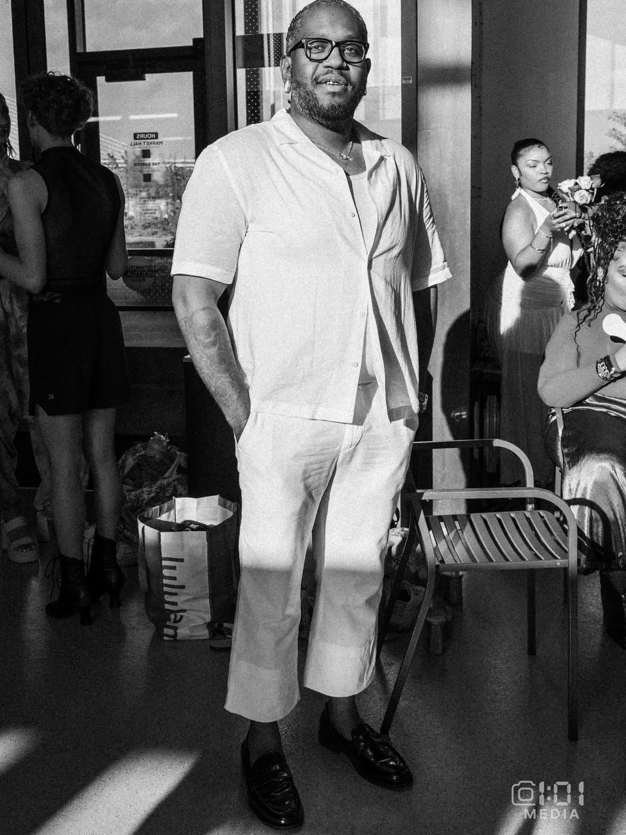 Man with glasses and beard in light-colored shirt and pants standing indoors, with people and chairs in background.