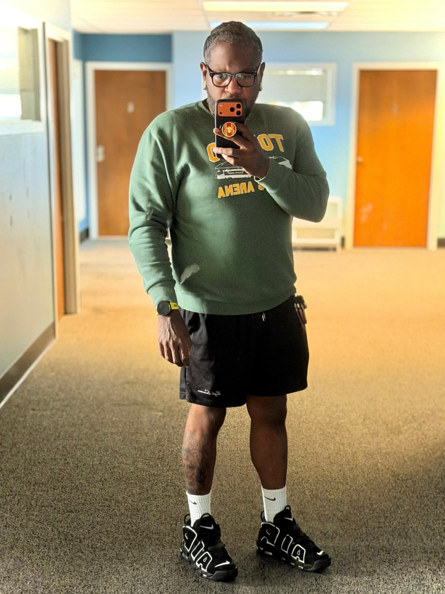 Person taking a mirror selfie in a room with blue walls and wooden doors, wearing a green sweatshirt, black shorts, and sneakers.