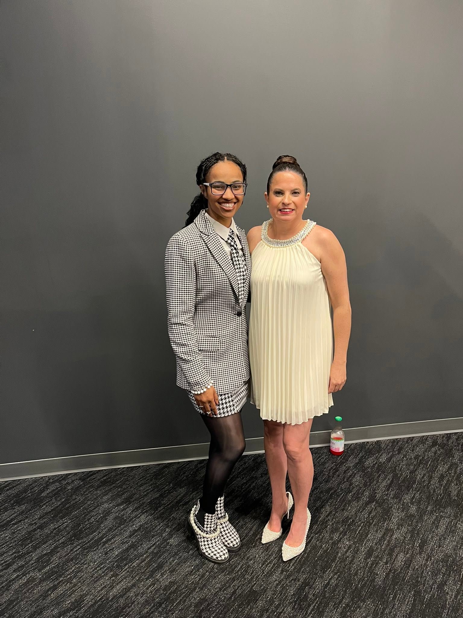 Two women standing side by side against a plain gray wall, smiling, one in a checkered blazer and the other in a cream dress.