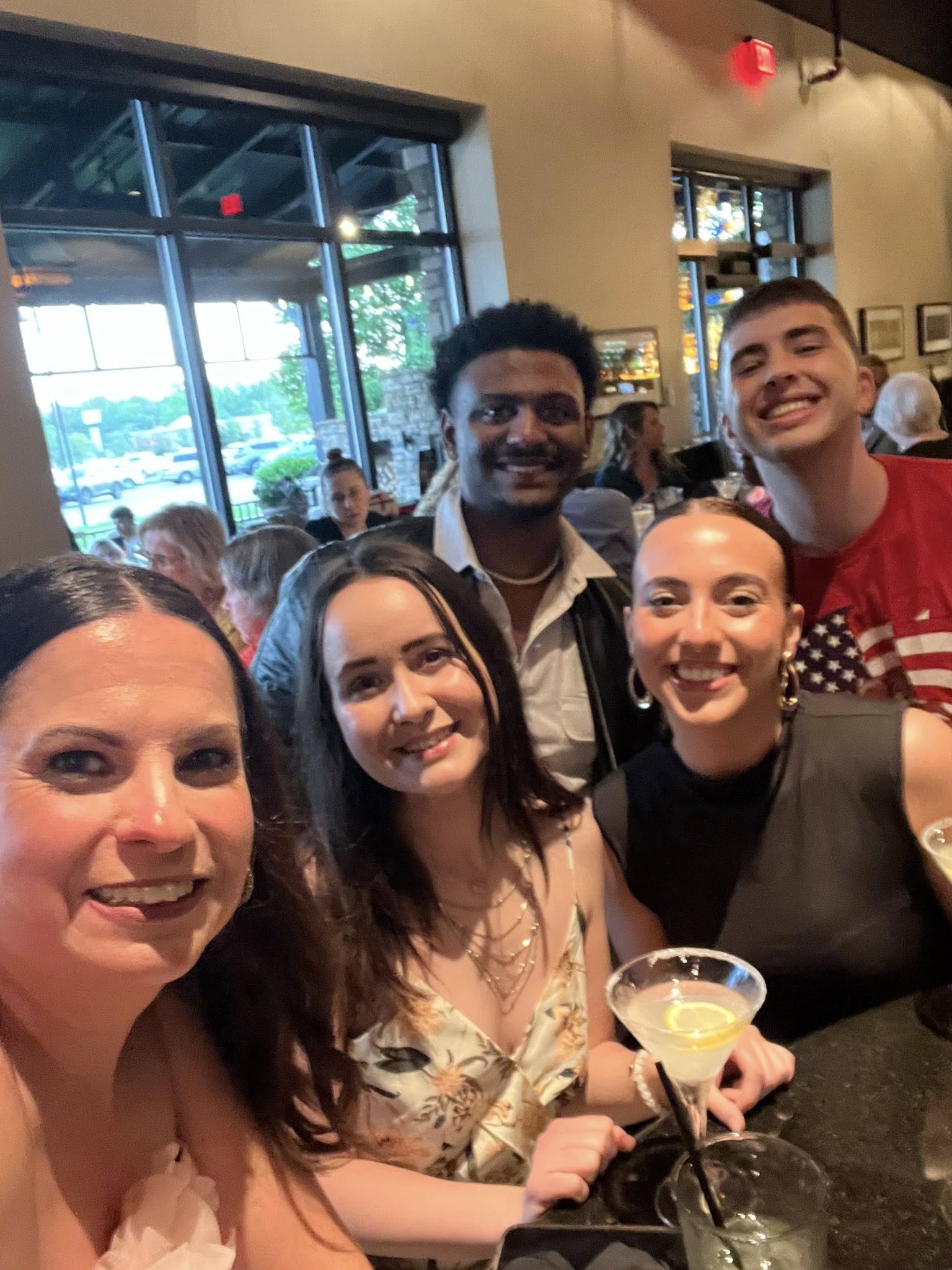 Group of five smiling people gathered inside a cafe or restaurant, with large windows and a bar in the background.