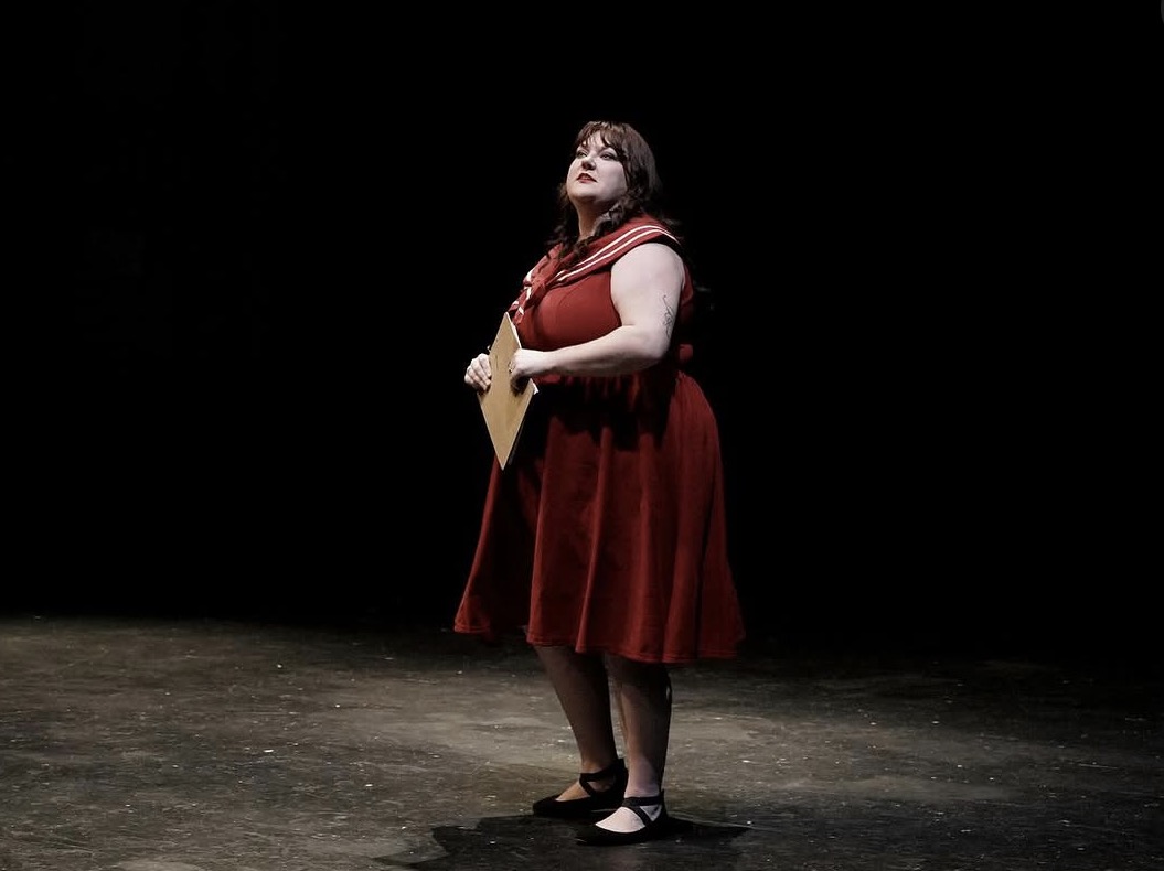 Woman standing on stage holding a guitar, wearing a red dress, with a black background.
