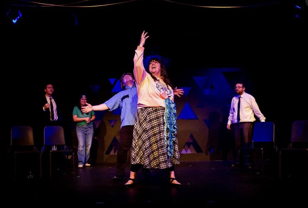 Group of five people performing on stage with a woman in front raising her arm, dark background, stage lights, chairs behind.