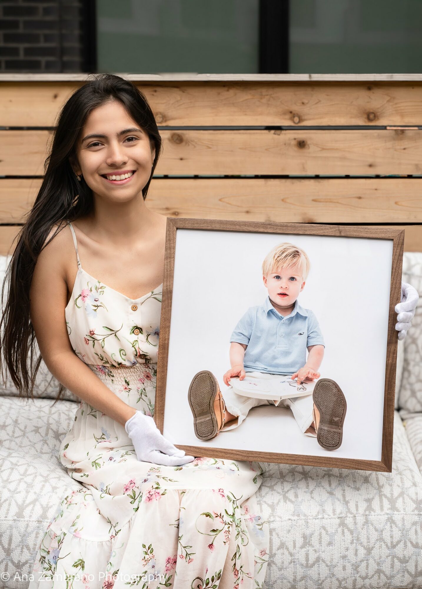 Young woman with long dark hair smiling, holding a framed photo of a child sitting on the ground, outdoors.
