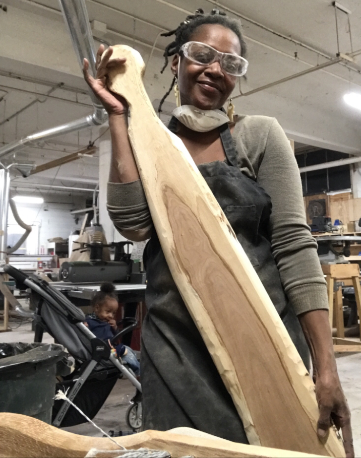 Woman in workshop holding a large wooden piece, wearing safety glasses and apron, smiling at the camera.