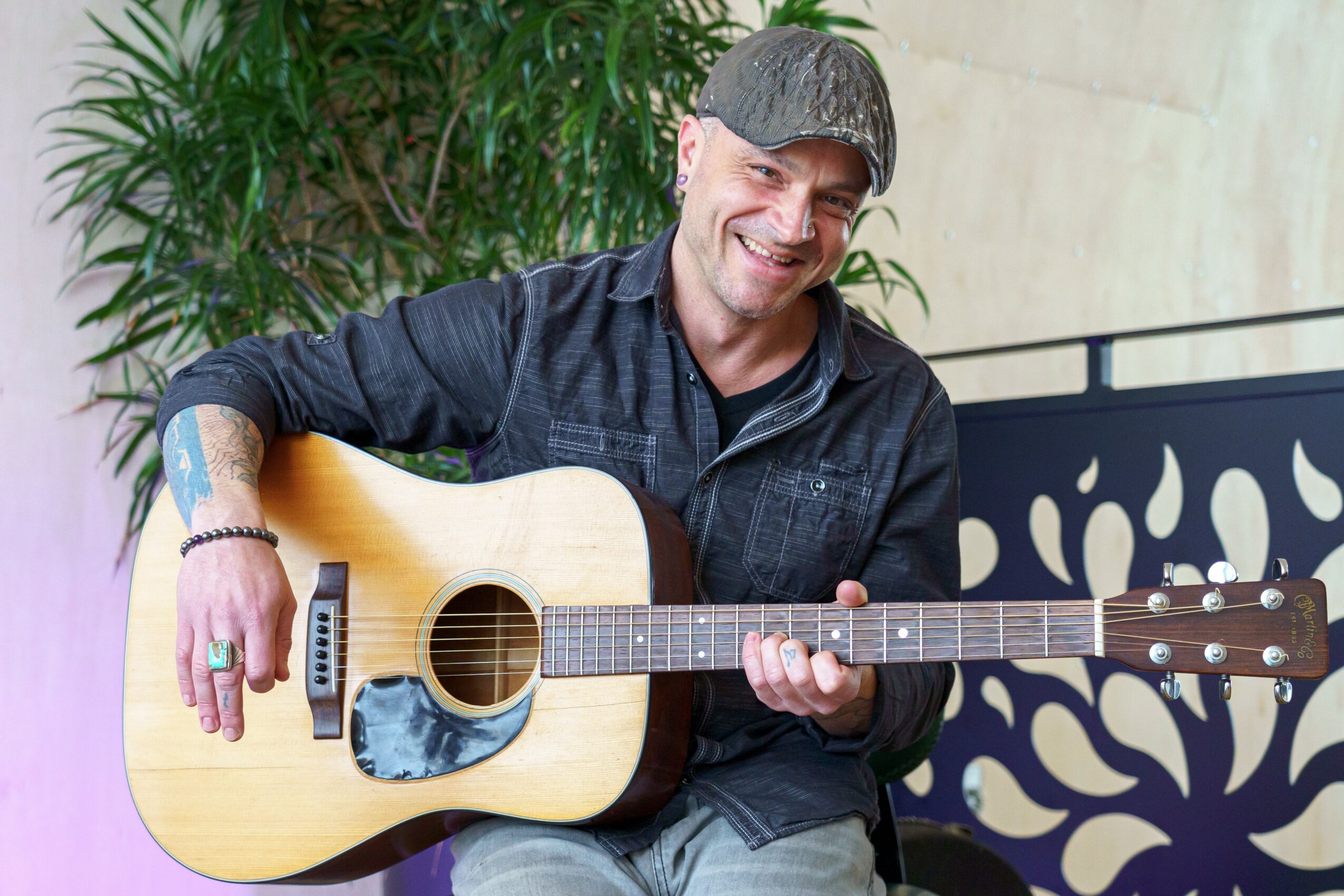Man smiling and playing an acoustic guitar indoors with a plant in the background.
