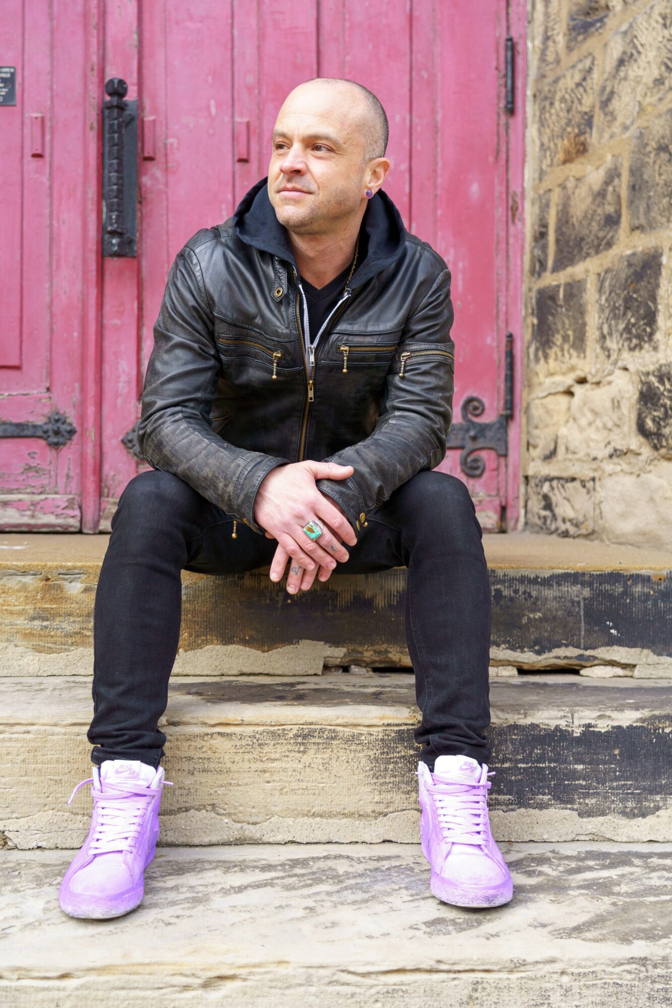 Bald man in black leather jacket and purple sneakers sitting on stone steps in front of a pink door and stone wall.