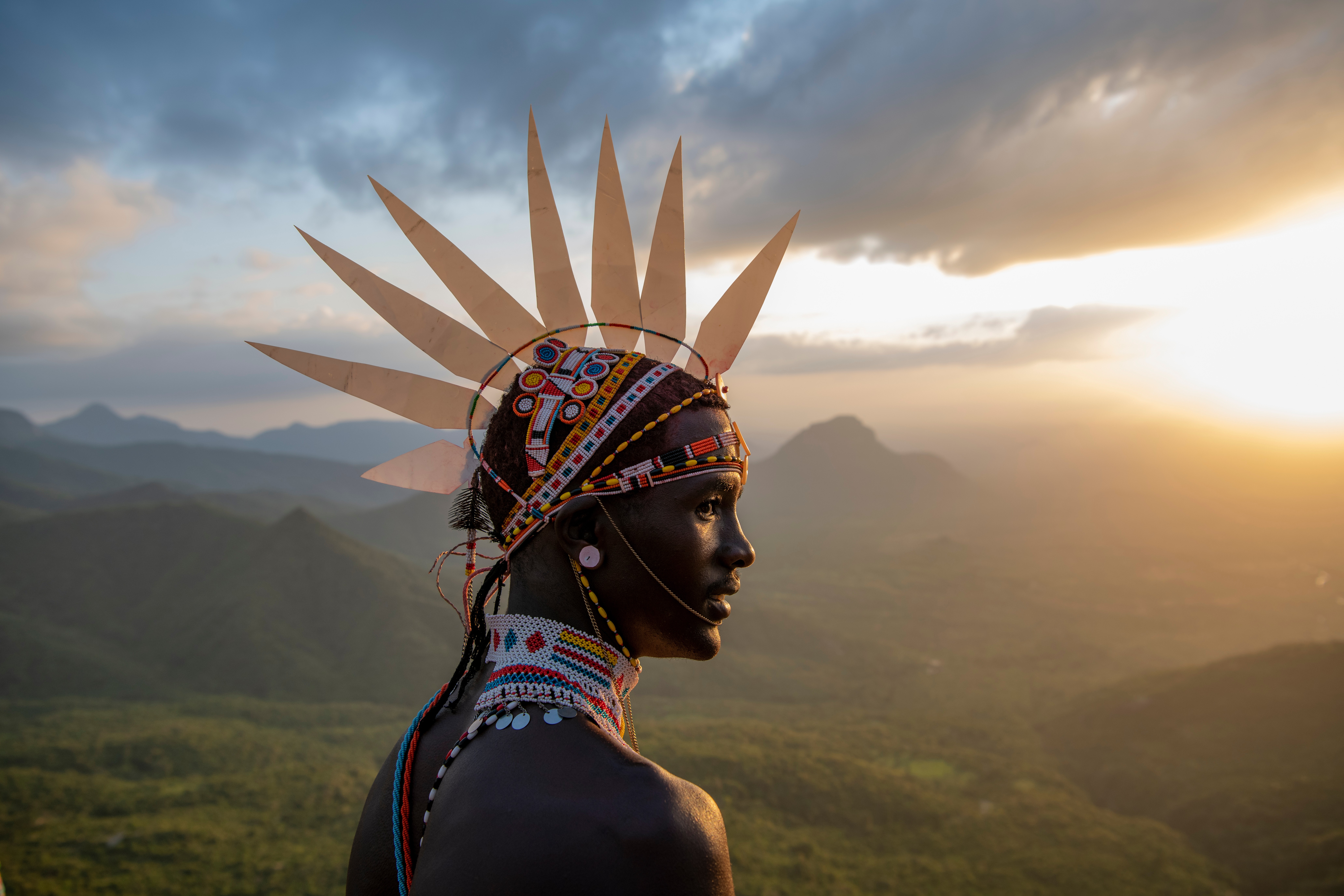 Profile of a person wearing a colorful headdress with pointed decorations, against a landscape at sunset.