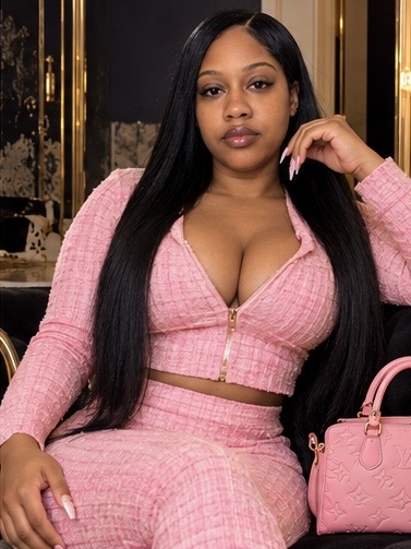 Young woman with long black hair, wearing pink outfit, sitting indoors with a pink handbag nearby.
