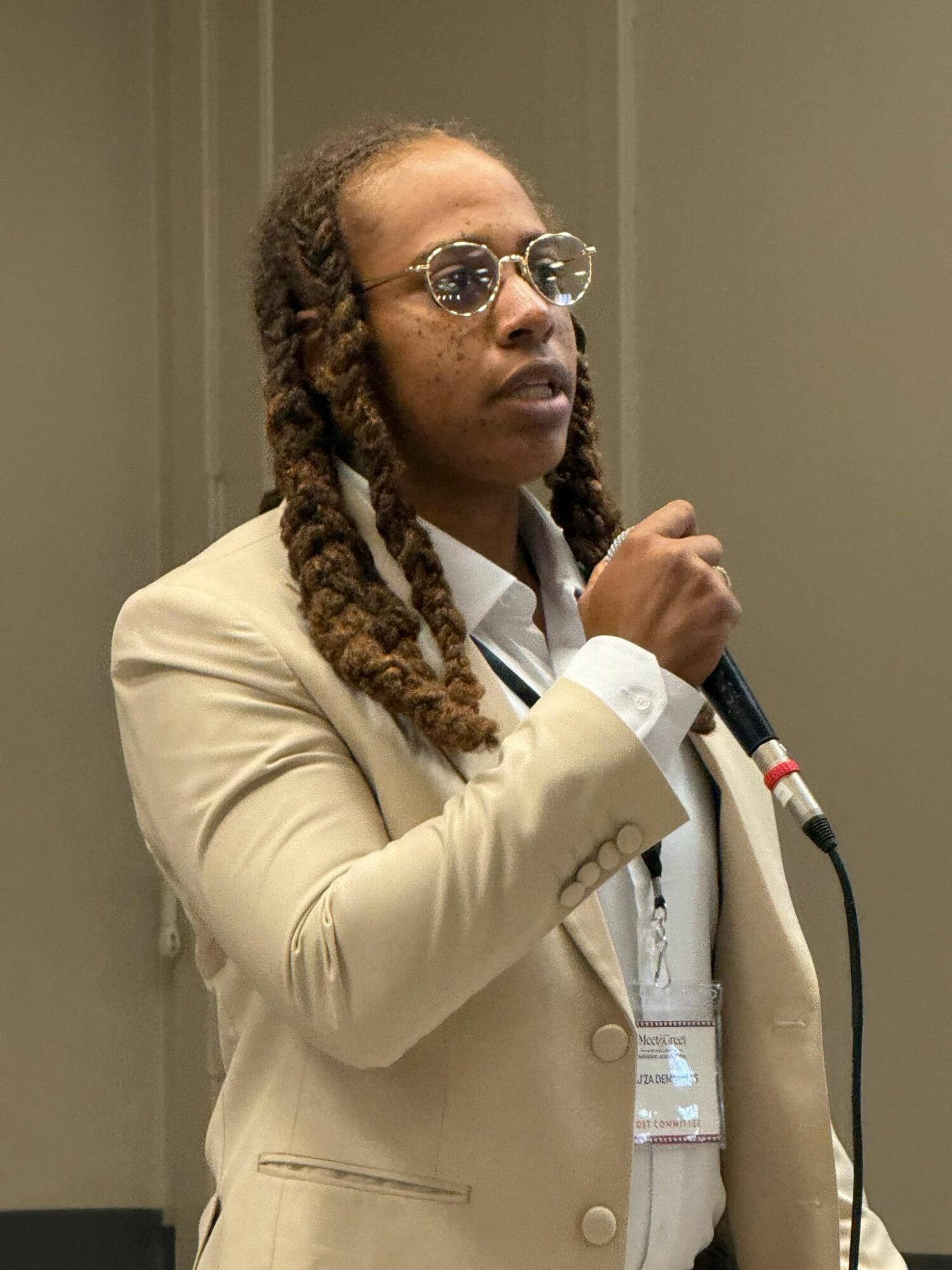 Woman with glasses and braided hair in a beige blazer holding a microphone, standing indoors.