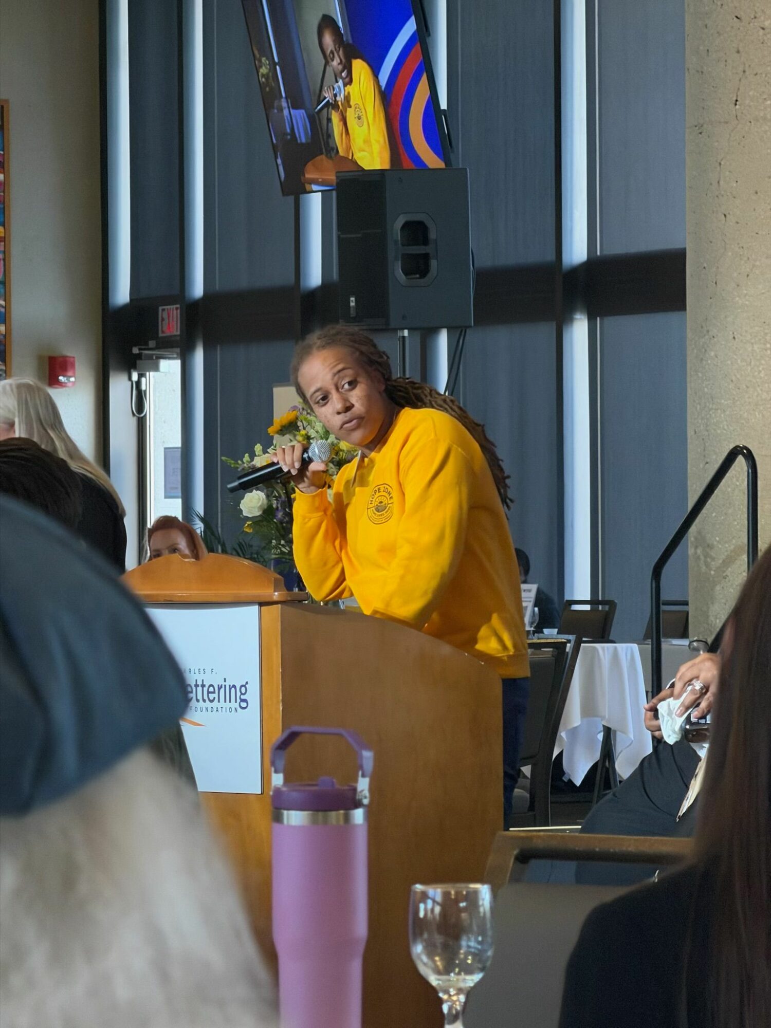Woman in yellow jacket speaking at a podium during a presentation or event, with a microphone and flowers, in a modern room.