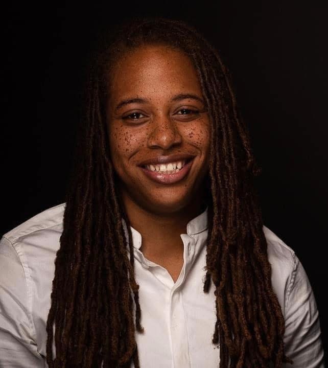Portrait of a smiling woman with long dreadlocks wearing a white shirt, dark background.