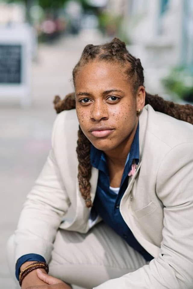 Woman with braided hair wearing a light-colored blazer and blue shirt, sitting outdoors with a blurred background.