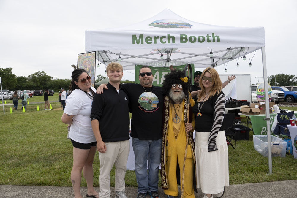 Group of five people standing in front of a white tent labeled 'Merch Booth' outdoors, some wearing costumes and sunglasses.