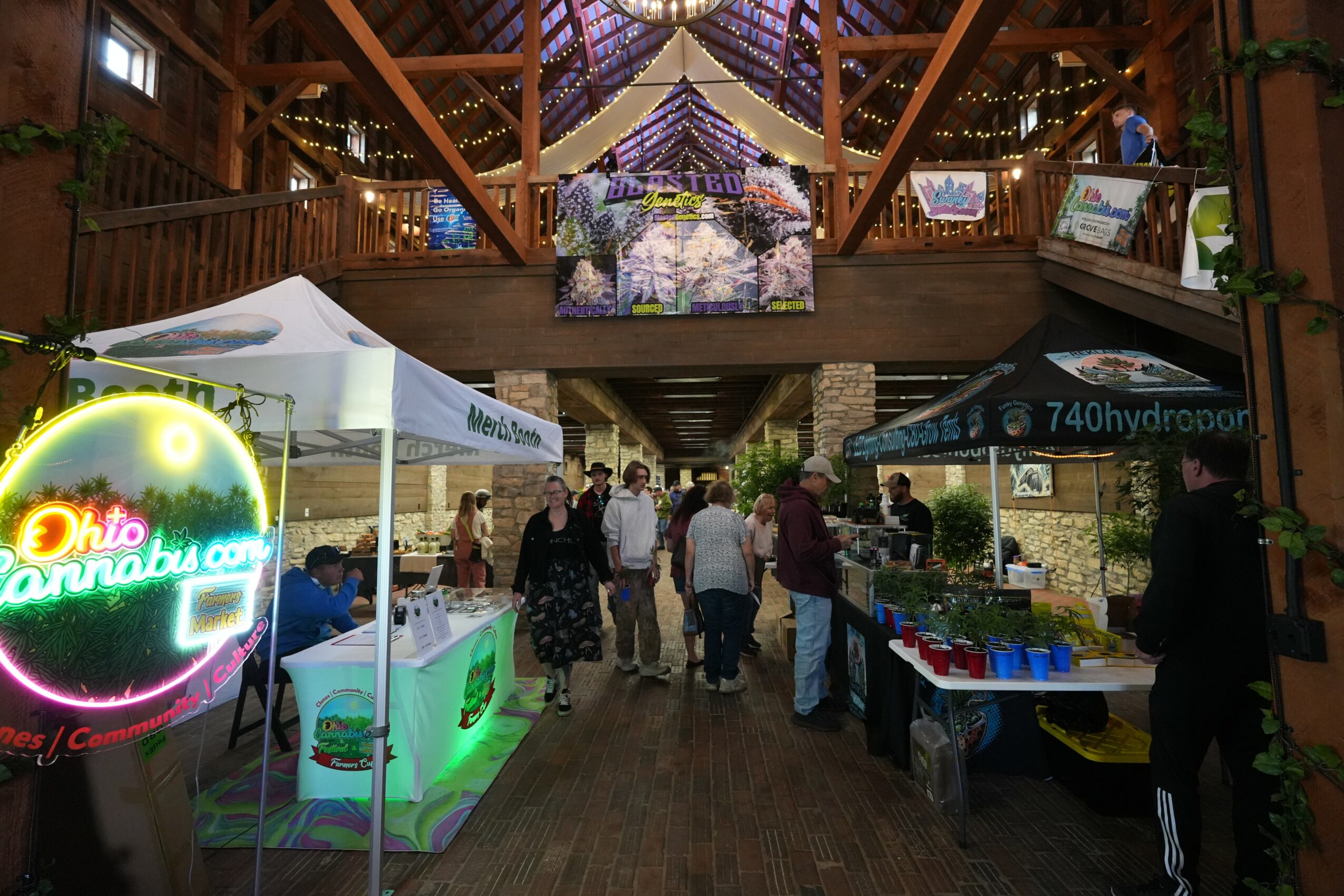 Indoor event with booths and people, decorated with a large floral display overhead, and a neon sign on the left.