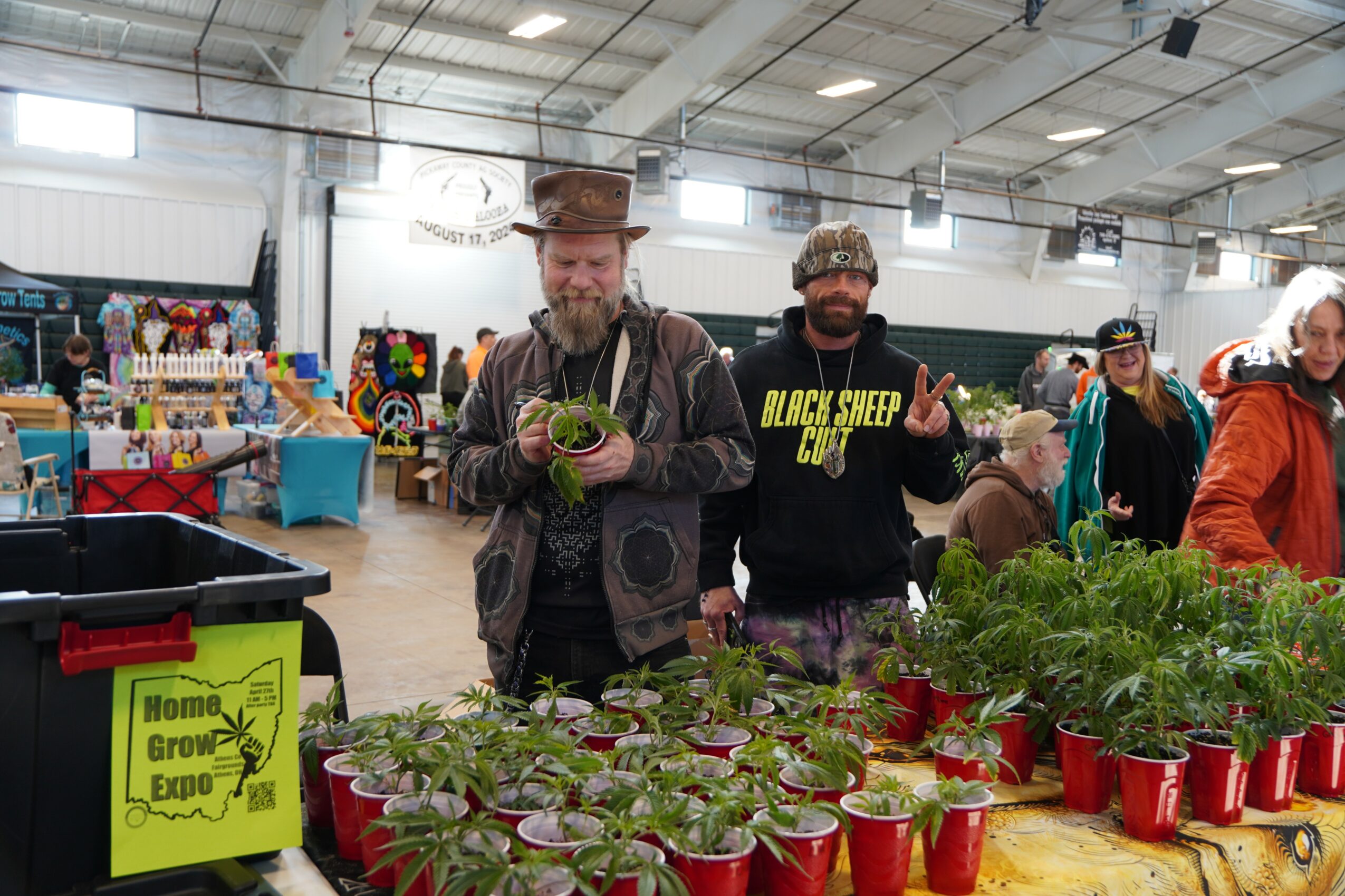 Two men at a market stall with plants and seedlings, one holding a small plant, in an indoor event space.
