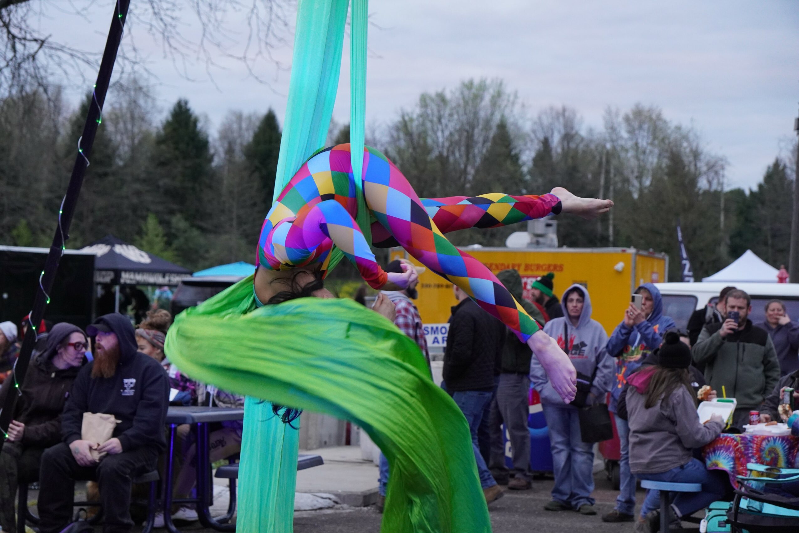 Colorful performer in a striped costume with long limbs, performing at an outdoor event with a crowd and trees in the background.