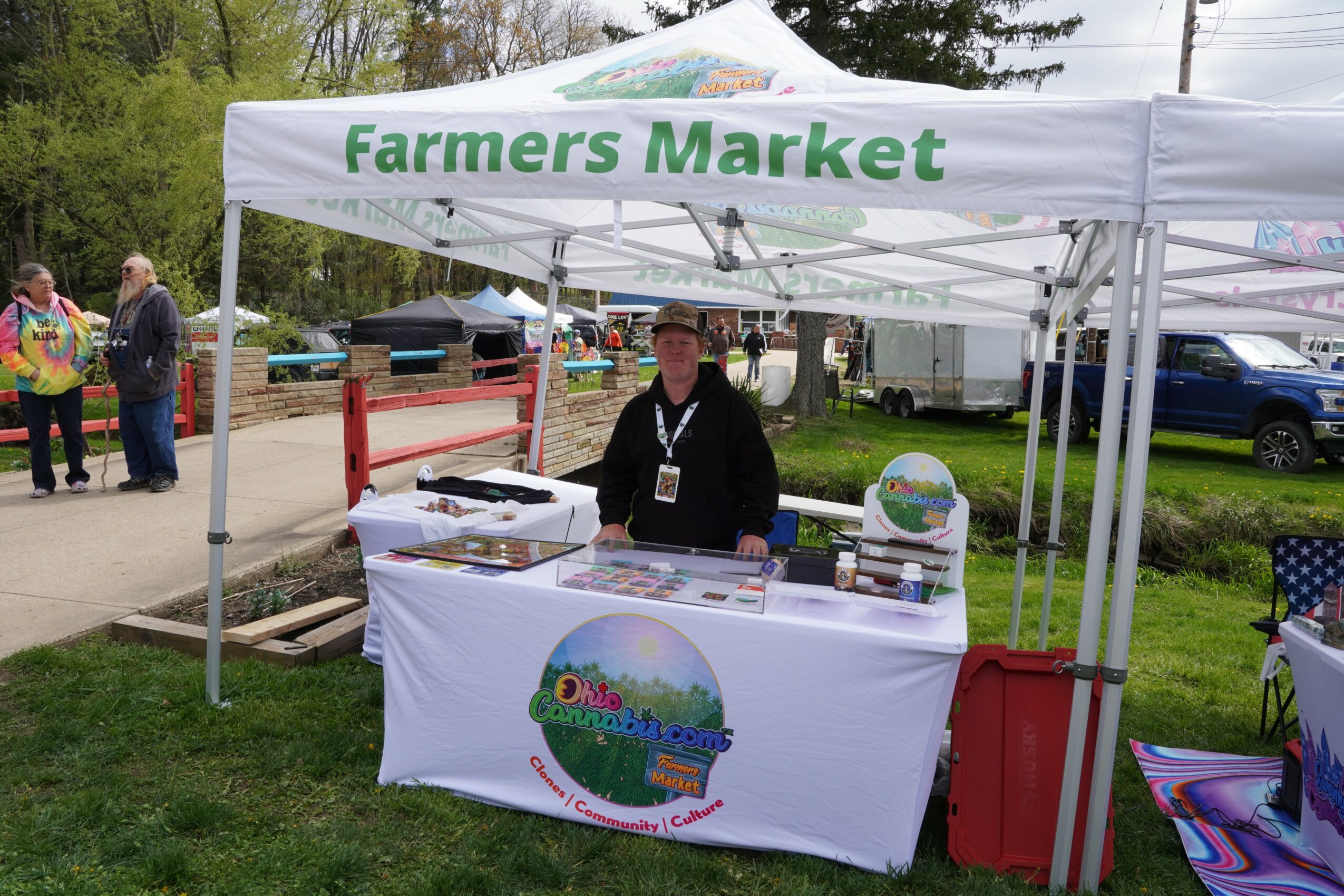 Person standing behind a Farmers Market booth with a white canopy, outdoor setting, and people in the background.