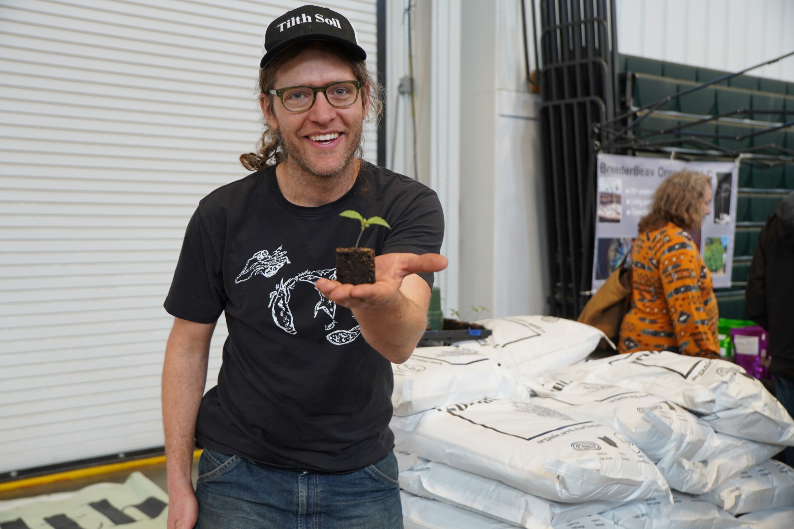 Person smiling and holding a small plant in a container, wearing glasses, a black cap, and a black t-shirt, standing indoors.