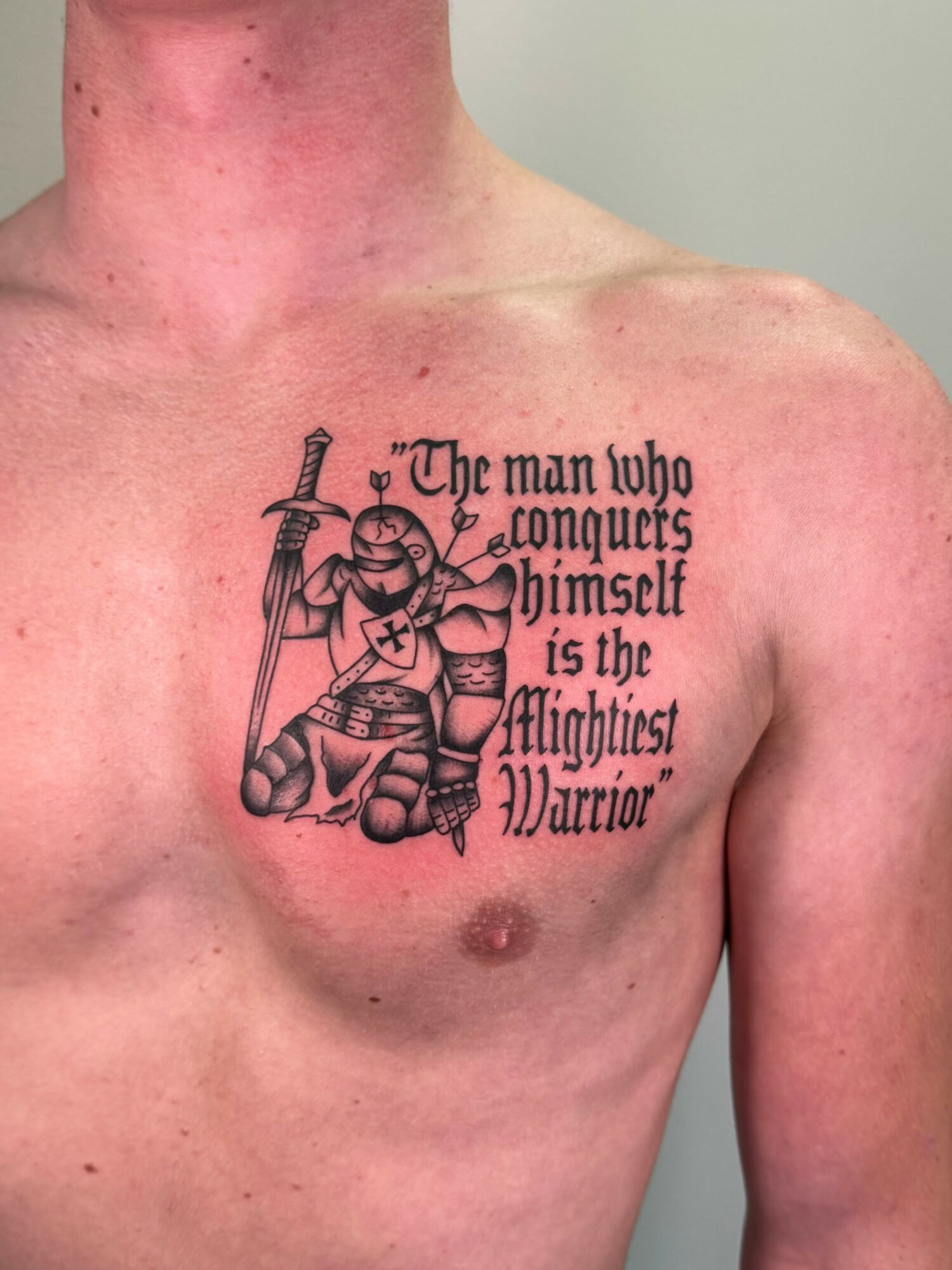 Tattoo of a warrior with a sword and shield on a person's chest, with quote about conquering oneself.