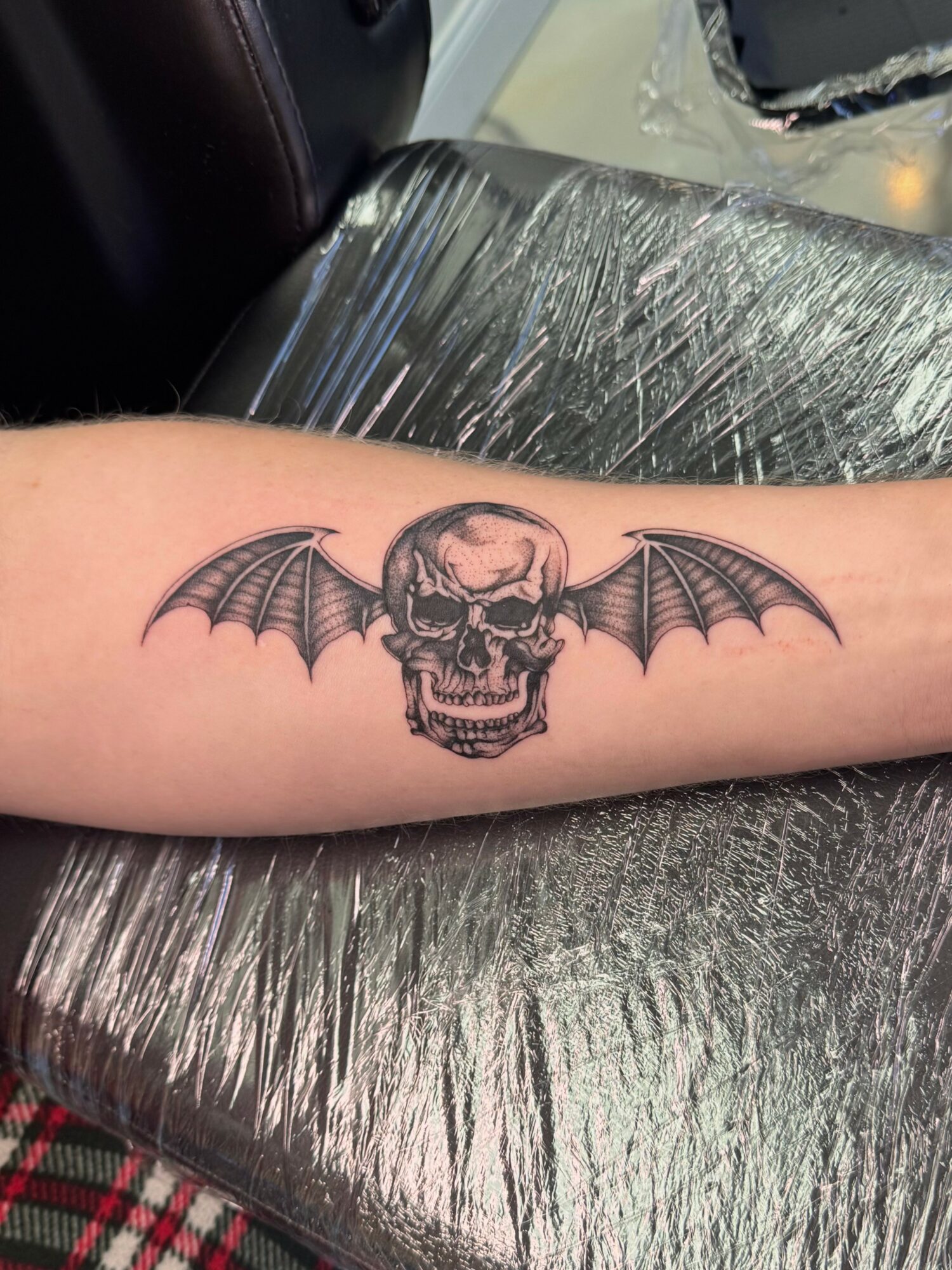 Tattoo of a skull with bat wings on a person's forearm, black ink, detailed design.