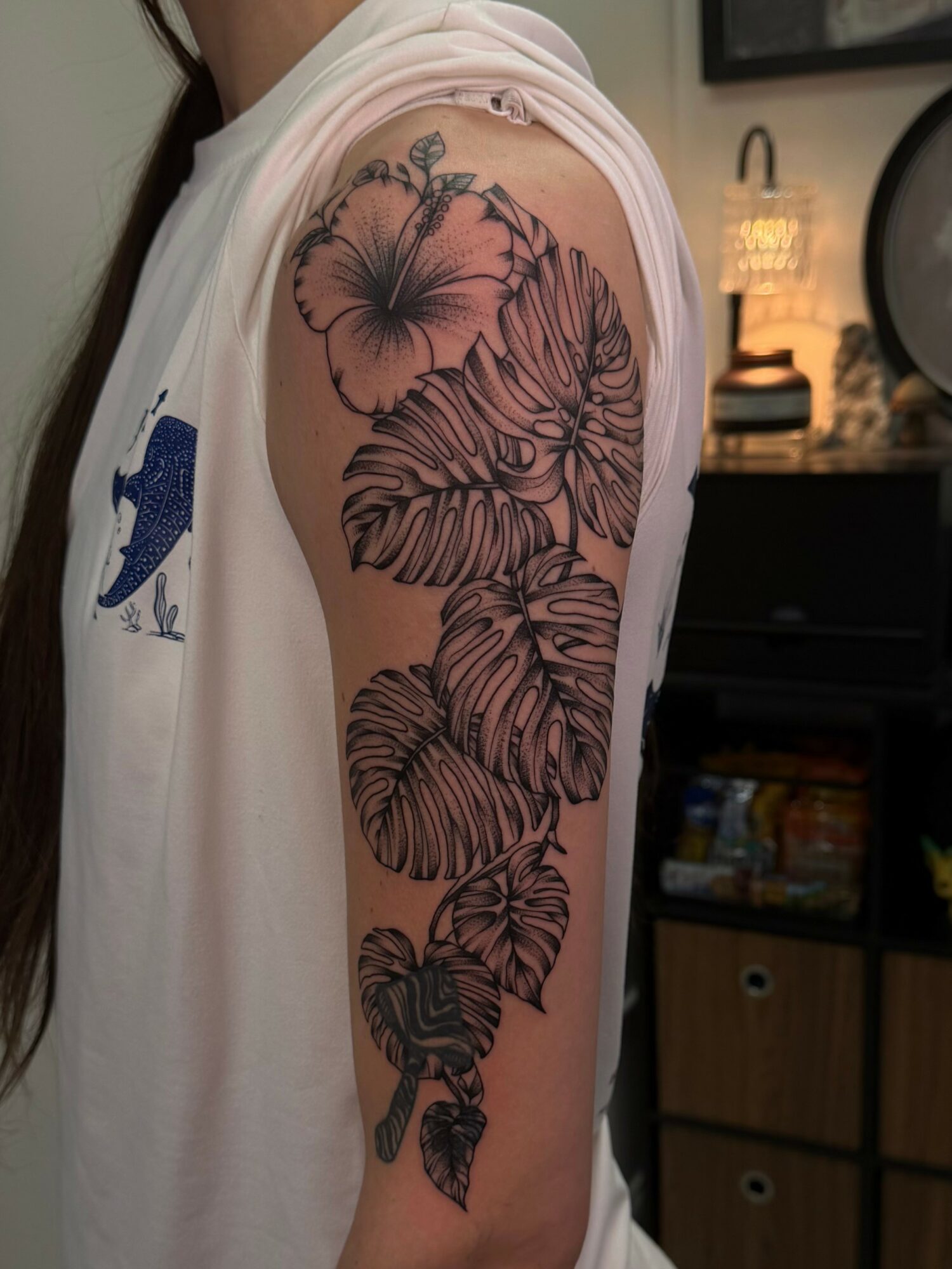 Tattoo of large tropical leaves on person's upper arm, with a background of a room with a lamp and shelves.
