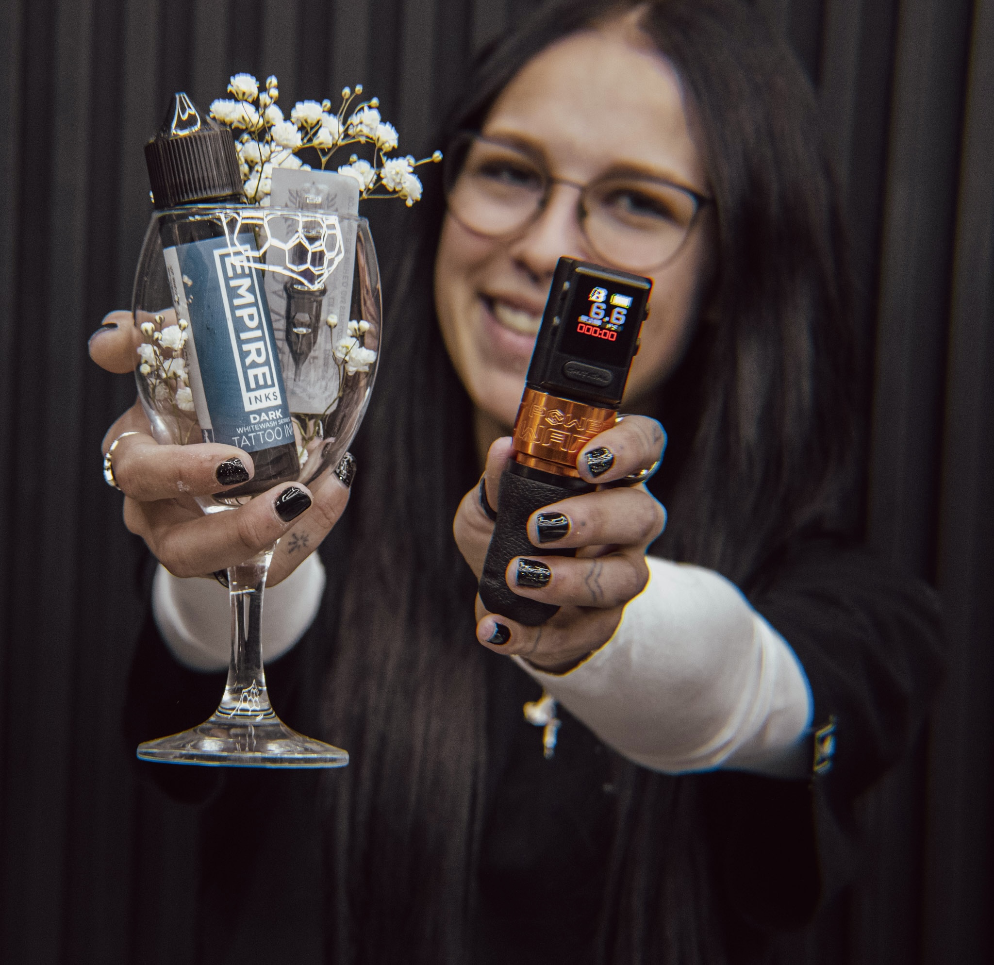 Woman smiling, holding a glass with flowers and a vape device, showing a small electronic device, against a dark background.