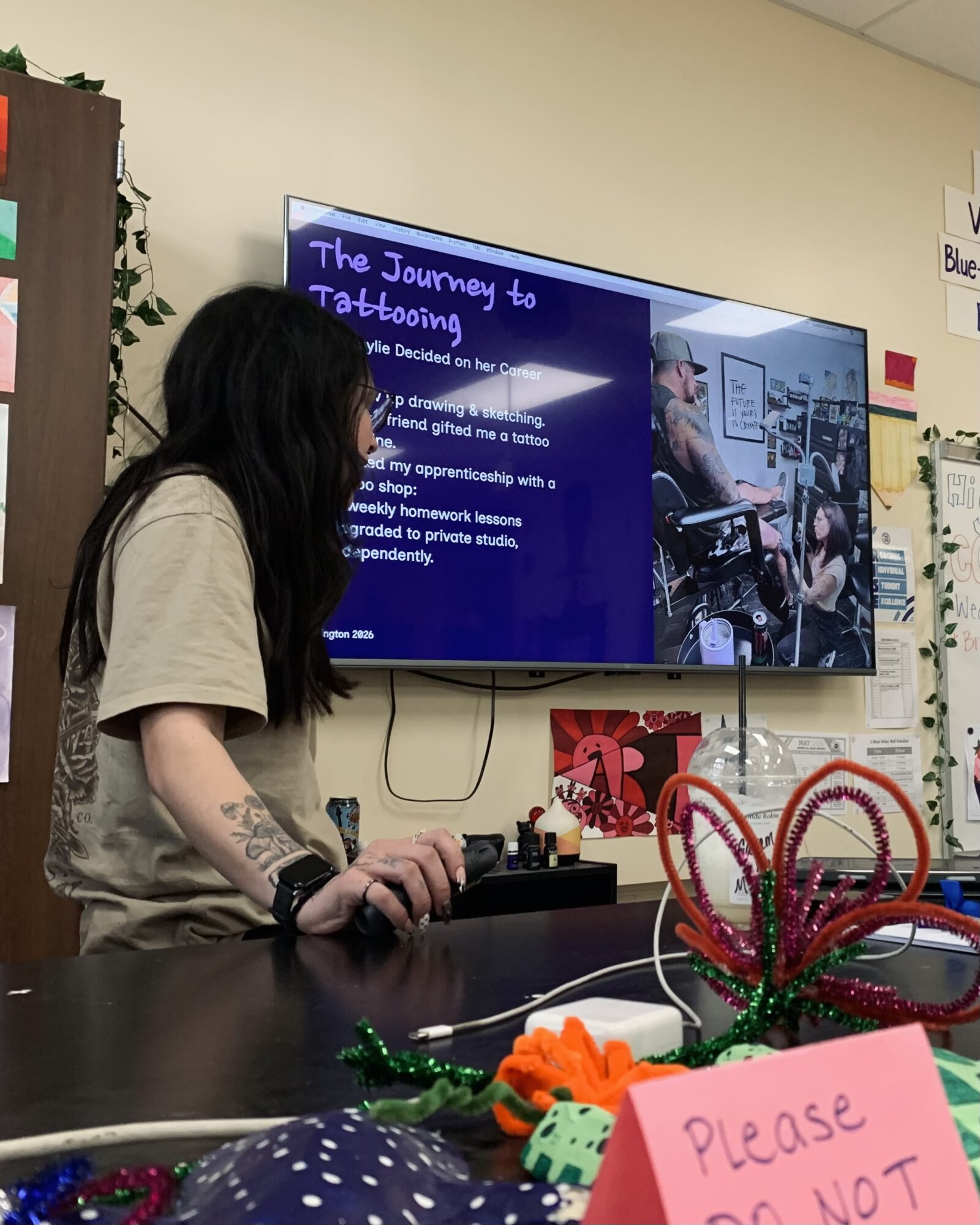 Person with long dark hair and tattoos on arm standing at a table with colorful craft materials, looking at a screen displaying a presentation about tattooing.