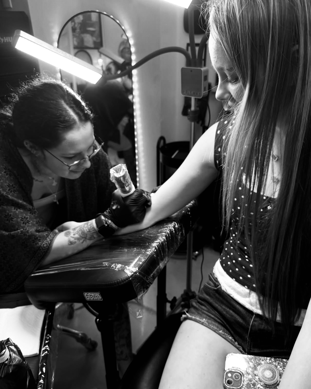 A person getting a tattoo on their arm by a tattoo artist in a studio, with equipment and lighting visible.