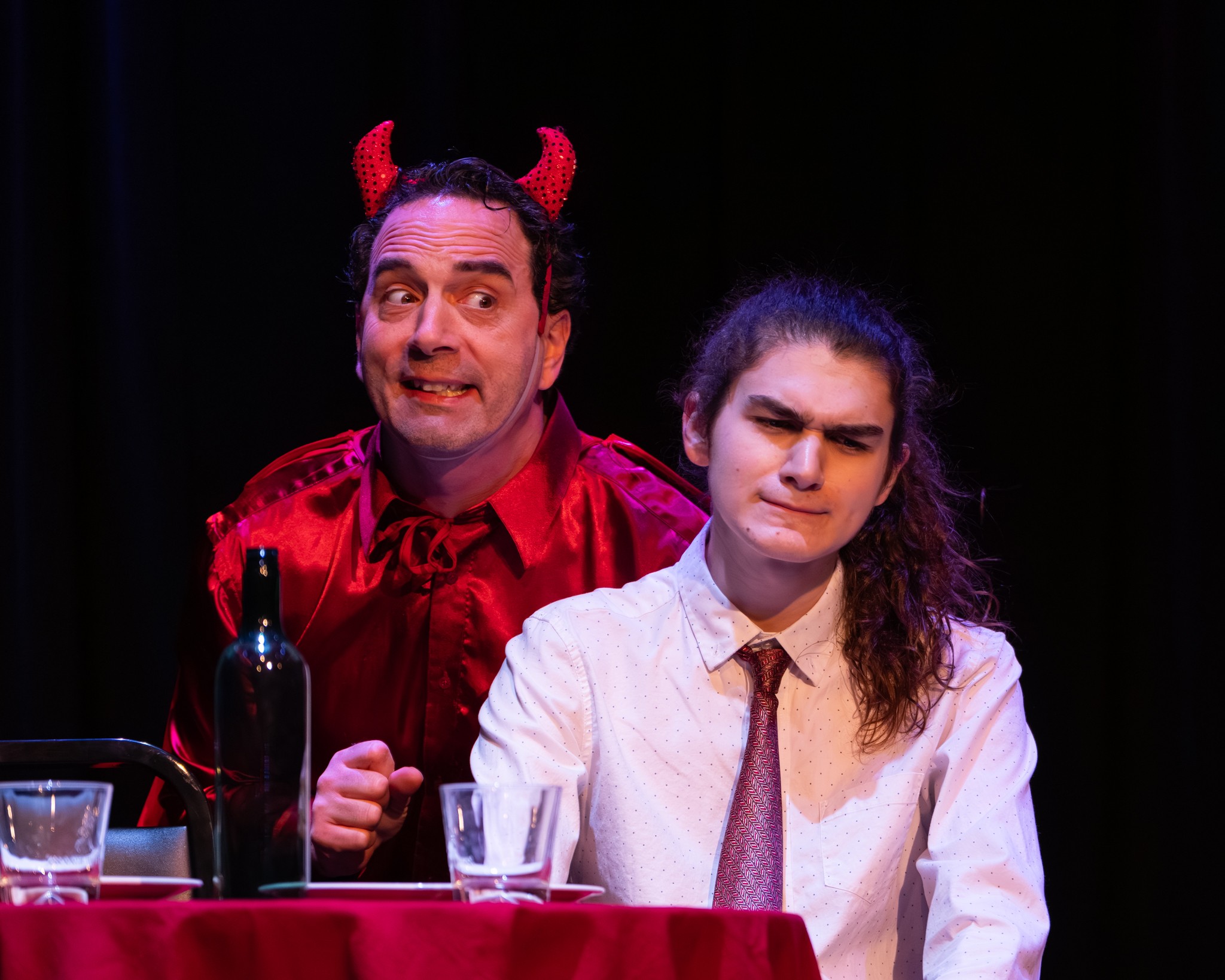 Two people sit at a table with drinks, one dressed as a devil with horns, the other with a serious expression.