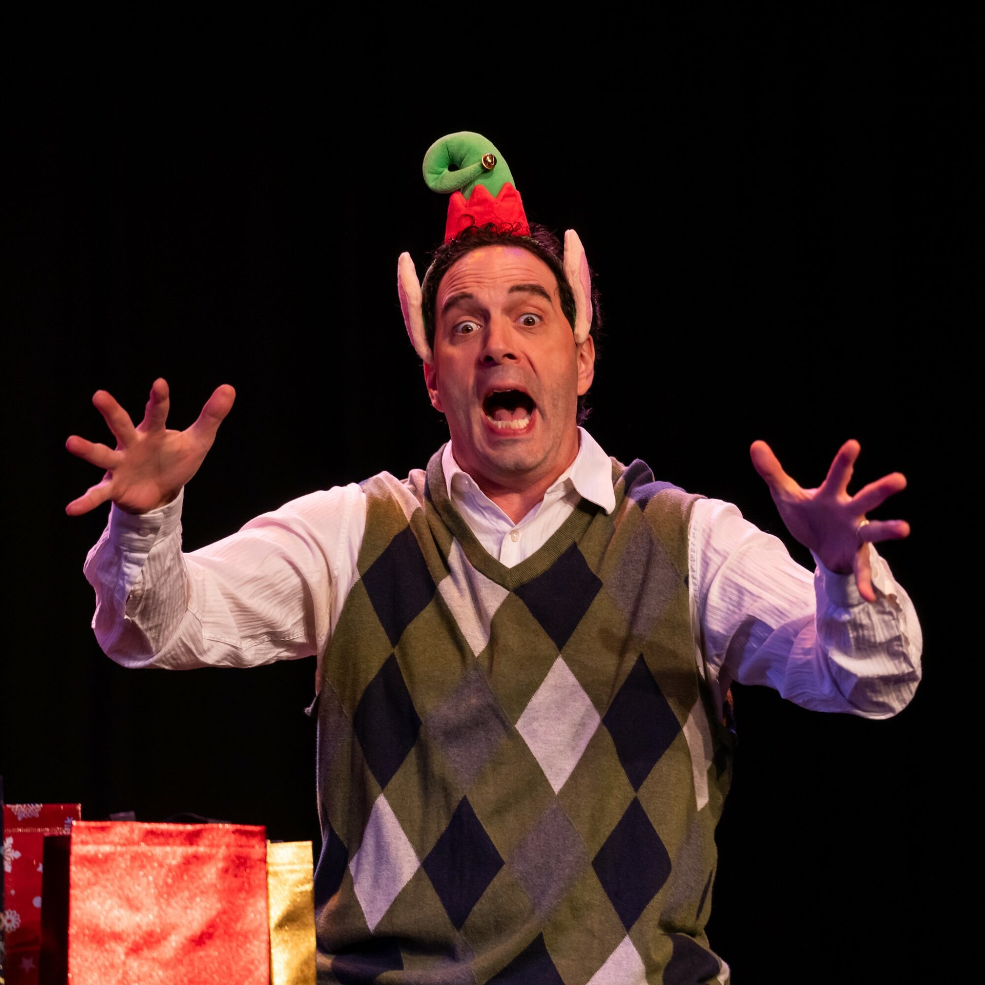Man with a surprised expression wearing a green and red bird hat, arms raised, on a dark stage.