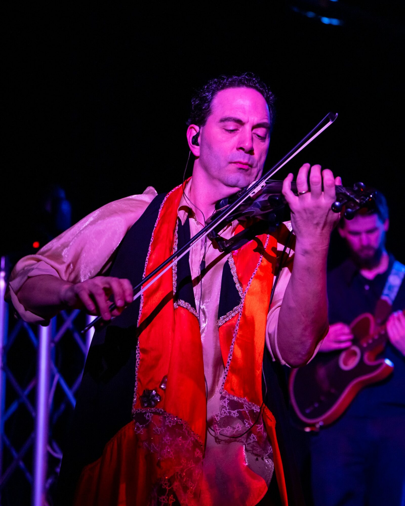 Man playing violin on stage with a guitarist in the background, dark setting, colorful lighting.