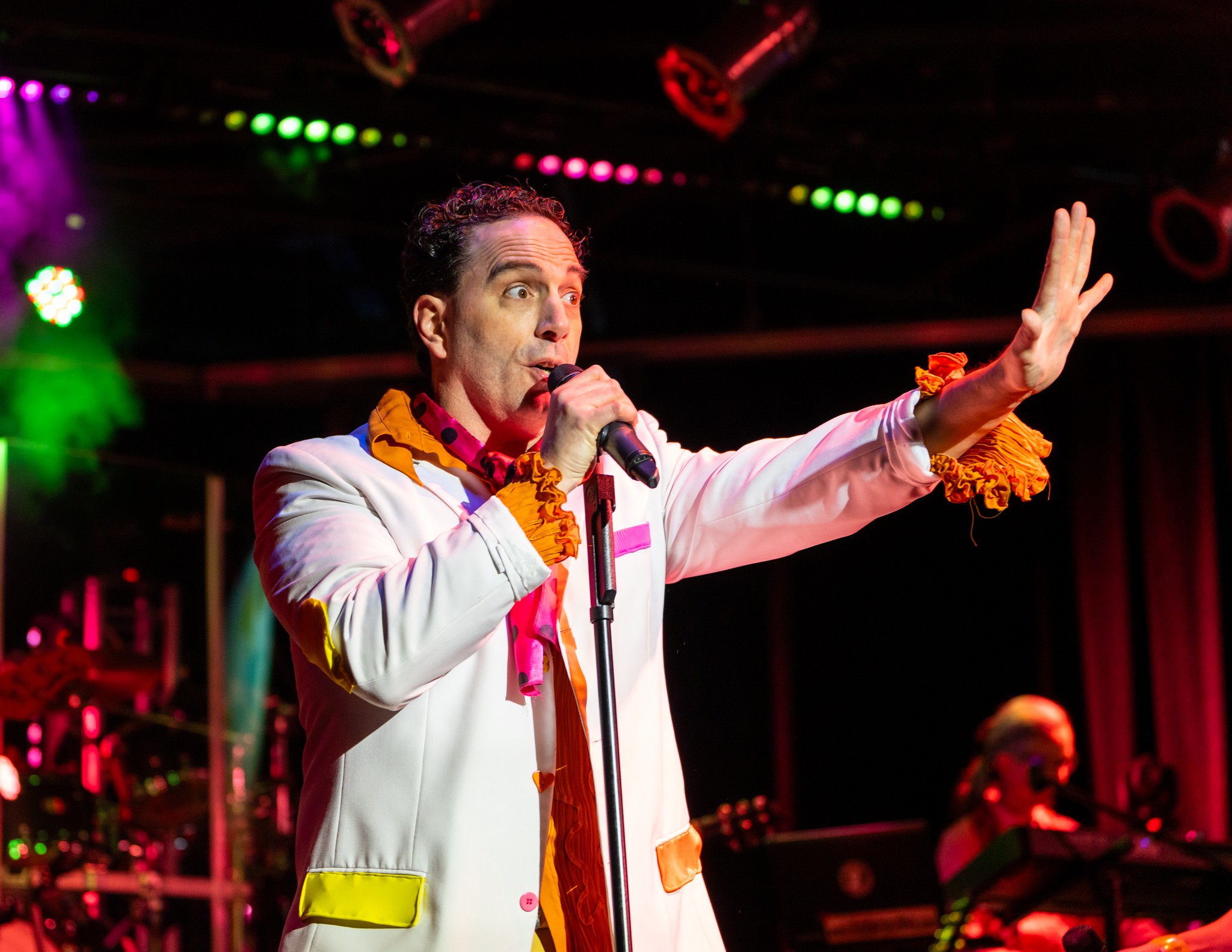 Performer singing on stage with colorful lights, wearing a white jacket and orange accessories, gesturing with one hand.
