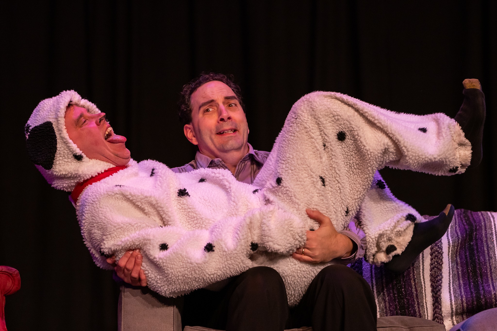 Person in a dog costume being held by a man on stage with a dark background.
