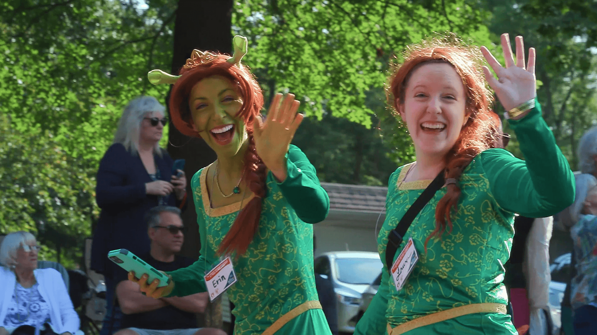 Two women in green costumes smiling and waving outdoors with trees and people in the background.