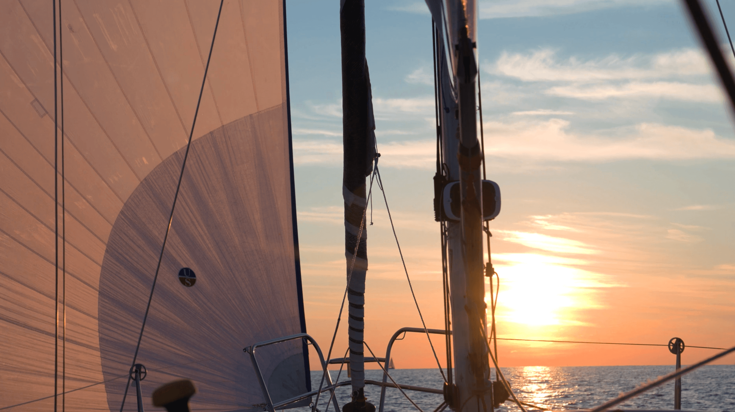Sailboat at sunset with sails and rigging visible, sun near horizon over water, sky partly cloudy.