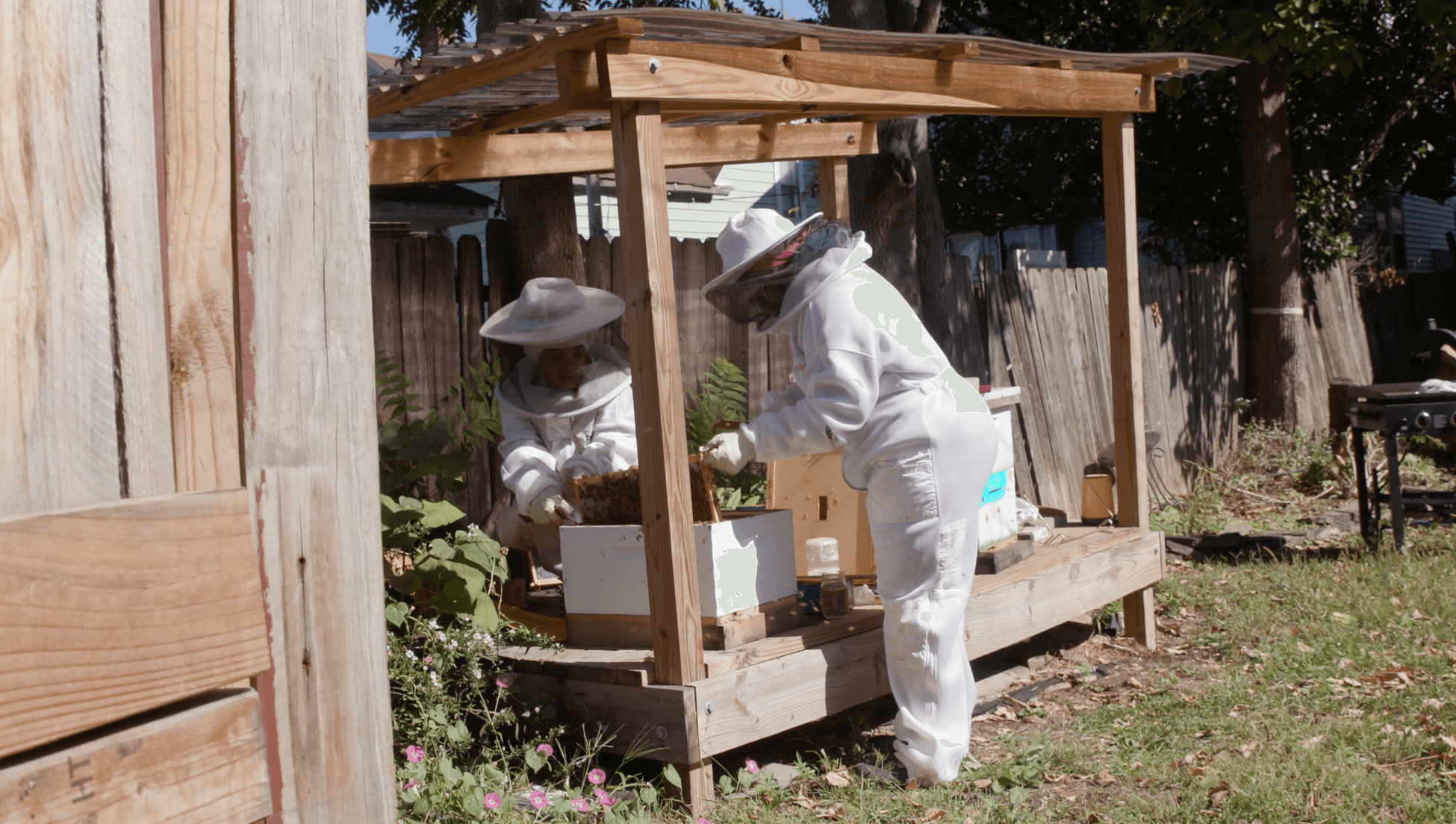 Two people in white protective suits and hats working at a wooden outdoor beekeeping hive, surrounded by grass and trees.