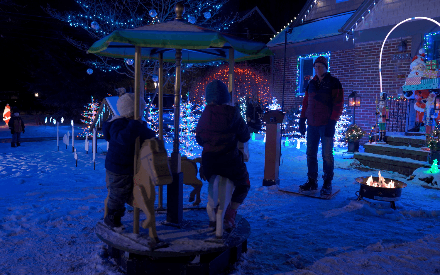 People gather outside decorated with blue and white lights, some near a small carousel, at night.