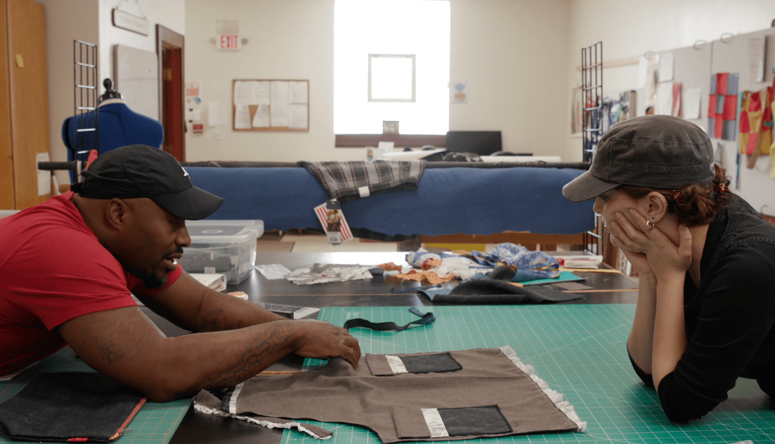 Two people sit at a table in a room, working on fabric or clothing projects, with supplies and materials around them.