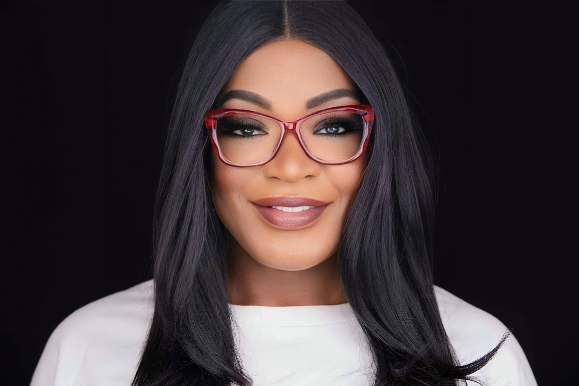 Woman with long black hair, red glasses, and makeup, smiling against a black background.