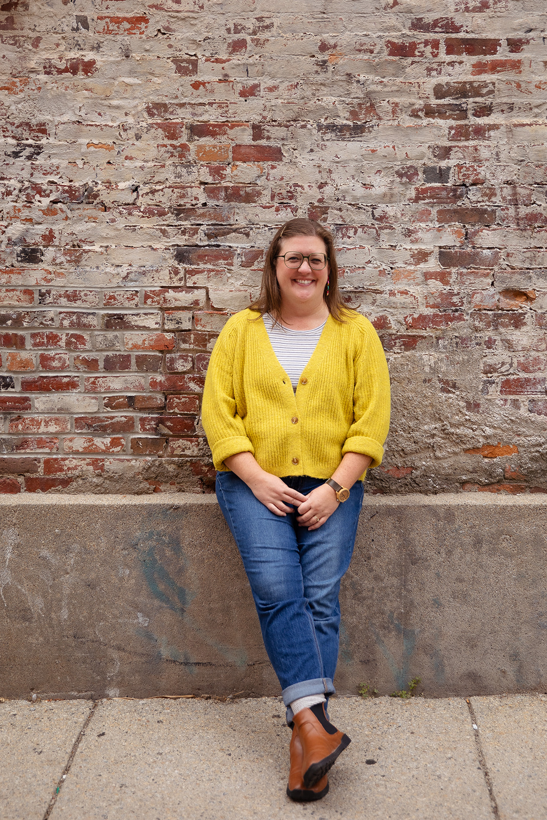 Woman with glasses and long hair, wearing a yellow cardigan and blue jeans, standing against a brick wall.