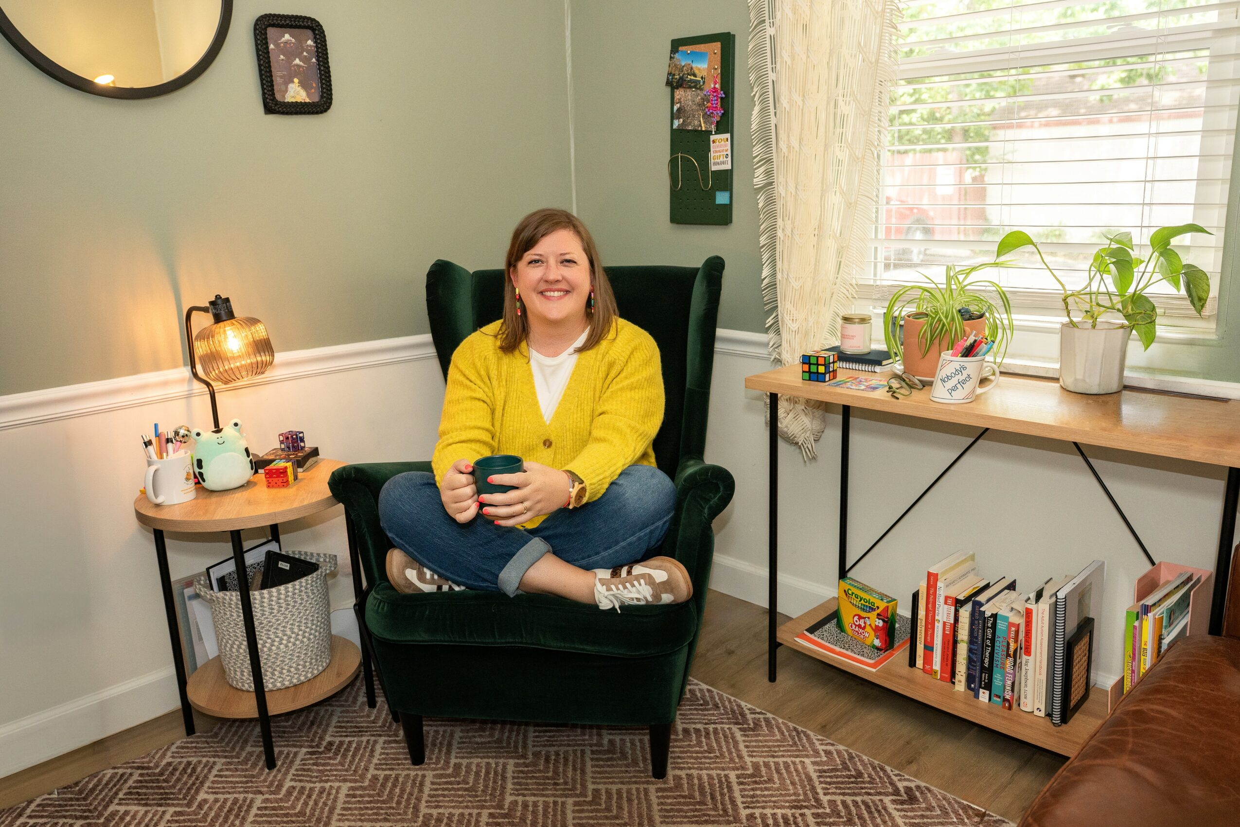 Smiling woman sitting in a green armchair holding a phone in a cozy room with a window, plants, and books.