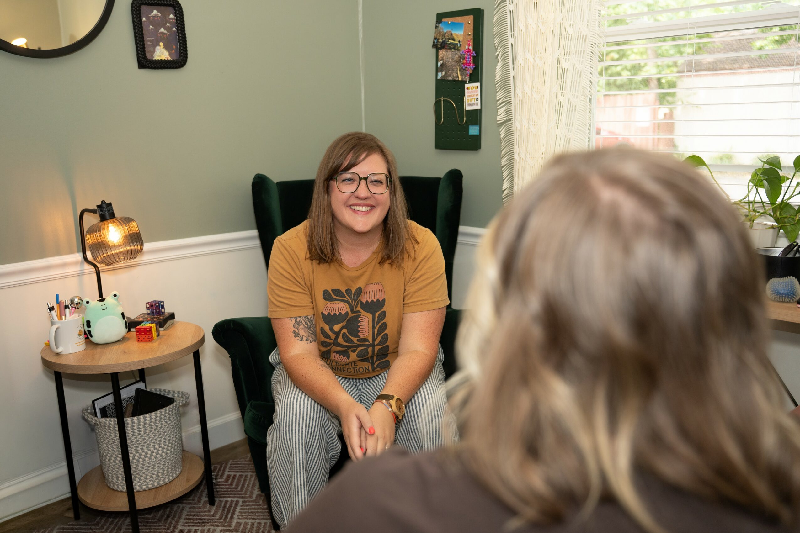 Young woman with glasses and a yellow shirt smiling, sitting in a green armchair in a room with a window and plants.