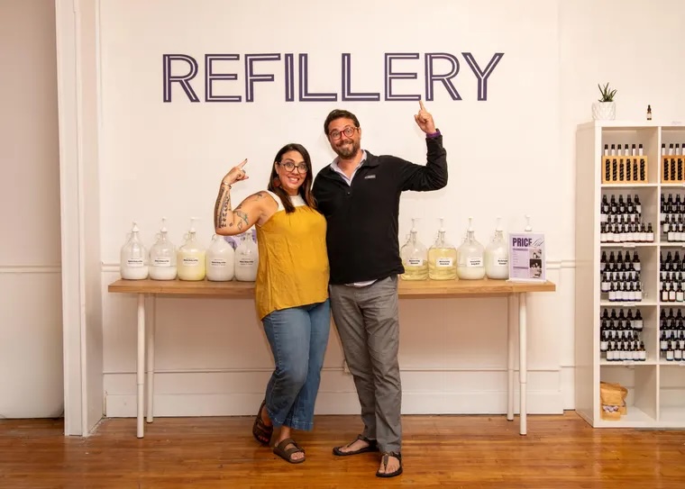 Two people stand in front of a table with jars, smiling and flexing their arms, in a store with a sign reading 'REFILLERY'.