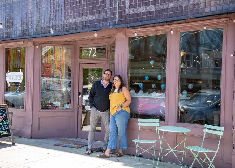 Two people stand outside a storefront with large windows and purple framing, smiling at the camera.