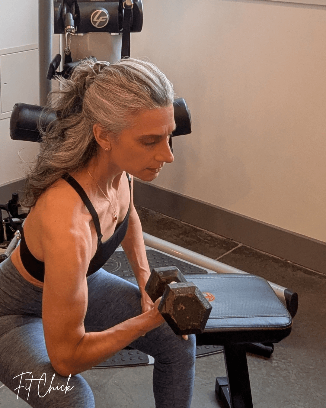 Woman with gray hair in a ponytail exercises with a dumbbell in a gym, sitting on a workout bench.