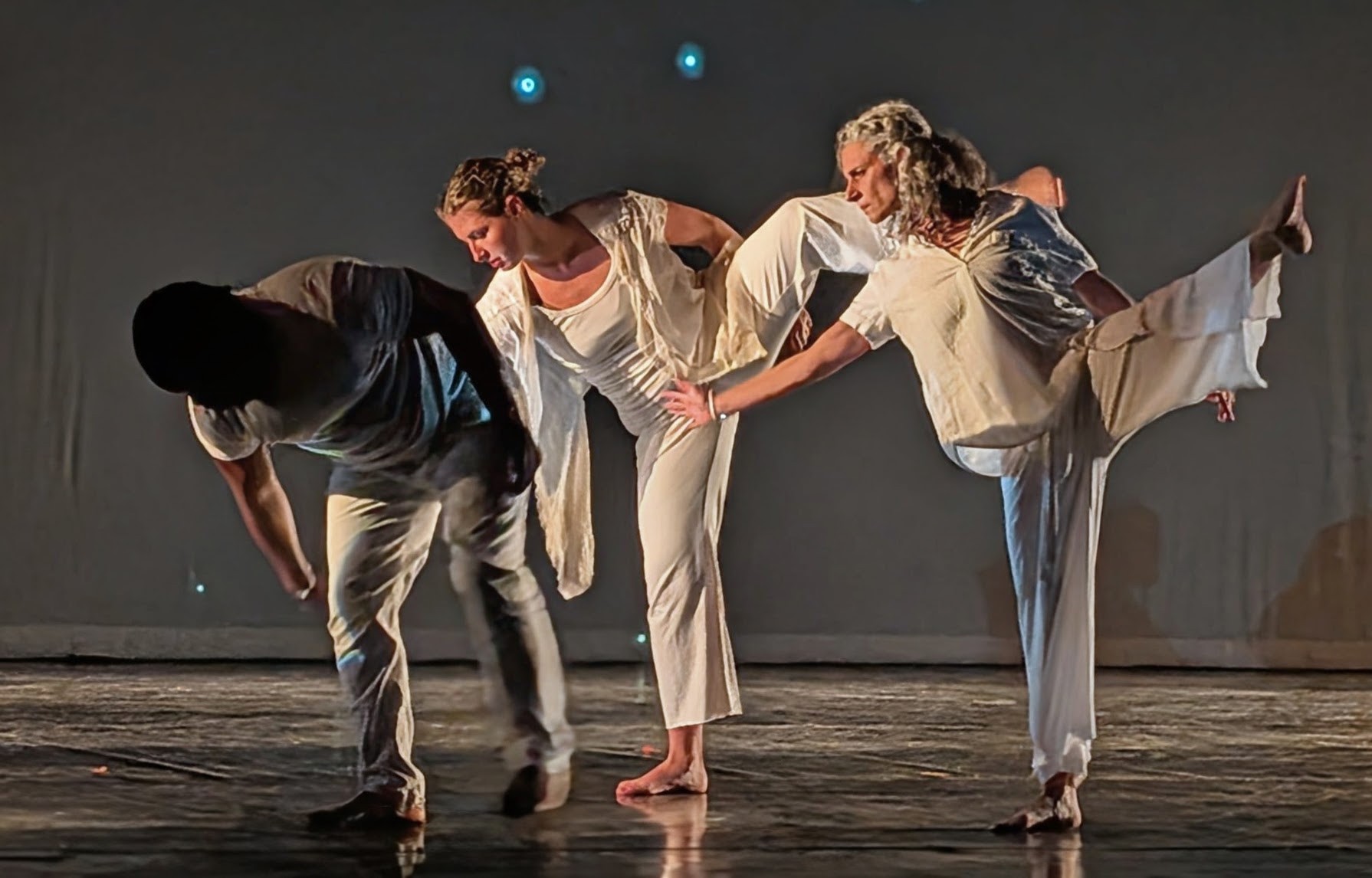 Four people perform a dance move on stage, holding hands and balancing on one leg, with a dark background.
